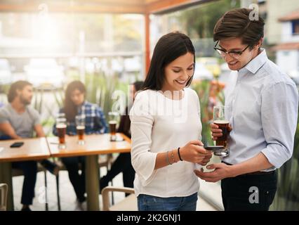 Wir sehen auf dem Bild so lustig aus. Aufnahme von zwei fröhlichen jungen Kollegen aus der Geschäftswelt, die Fotos auf einem Telefon draußen in einem Restaurant angeschaut haben. Stockfoto