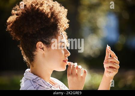 Es ist immer Zeit für Lippenstift. Kurzer Schuss einer attraktiven jungen Frau, die im Park einen Lippenstift aufgesetzt hat. Stockfoto