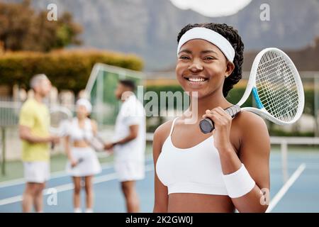 Ich bin bereit für das Spiel. Verkürztes Porträt einer attraktiven jungen Tennisspielerin, die draußen auf dem Platz mit ihrem Trainer und ihren Teamkollegen im Hintergrund steht. Stockfoto