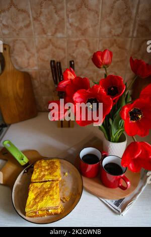 Rote Tasse mit heißem Kaffee im Hintergrund der Küche. Köstliche Toasts mit Ei liegen auf einer Pfanne. Vase mit roten Mohnblumen, Kaffeepause. Stockfoto