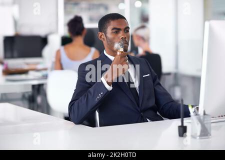 Fast so gut wie das echte... ein junger Geschäftsmann, der an seinem Arbeitsplatz eine elektronische Pfeife raucht. Stockfoto