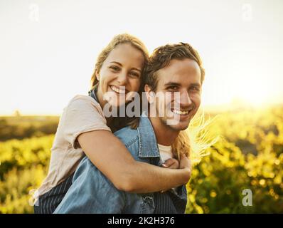 Wir haben immer Spaß draußen in der Natur. Aufnahme eines jungen Paares, das durch ihre Farm läuft, während es eine Huckepack-Fahrt mit einem Lächeln auf den Lippen hat. Stockfoto