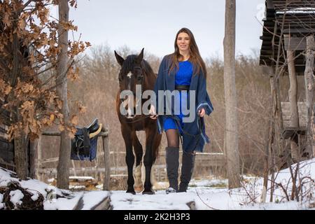 Ein Mädchen in blauem Kleid geht mit einem Pferd durch den Winterwald und alte Holzruinen Stockfoto