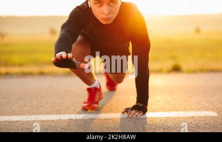 Entschlossener männlicher Athlet, der in der Crouch-Startposition steht und sich während des Trainings auf dem Land für das Laufen vorbereitet, während er die Kamera anschaut Stockfoto