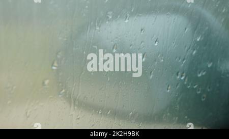 Verschwommener Regen fällt auf ein Autofenster mit dem Spiegel im Hintergrund. Nahaufnahme Stockfoto