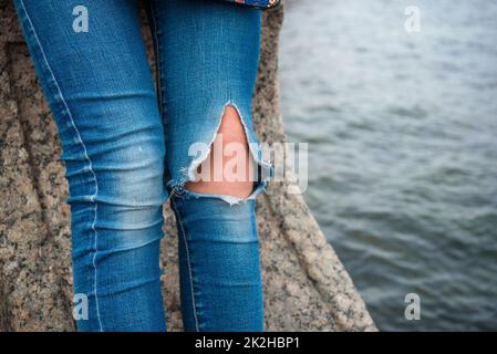 Frauenbeine in Jeans, die am Knie gerissen sind, sitzen auf dem Pier gegen das Wasser Stockfoto