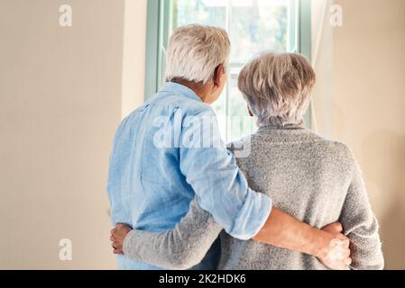 Erinnerungen hinter ihnen, die Zukunft vor ihnen. Rückansicht eines liebevollen Seniorenpaares, das aus einem Fenster zu Hause zusammenschaut. Stockfoto