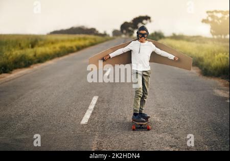 Dieser Skater weiß, wie er aufsteigen kann. Aufnahme eines Jungen, der vorgibt, mit einem Paar Flügel aus Pappe zu fliegen, während er draußen auf einem Skateboard reitet. Stockfoto