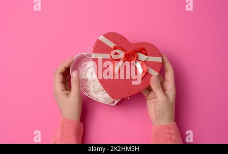 Zwei weibliche Hände halten eine rote herzförmige Box auf einem rosa Hintergrund. Festlicher Hintergrund Stockfoto