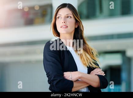 Sie hat eine Menge Gedanken im Kopf. Kurze Aufnahme einer attraktiven jungen Geschäftsfrau, die aufmerksam aussieht, während sie in einem modernen Büro mit verschränkten Armen steht. Stockfoto