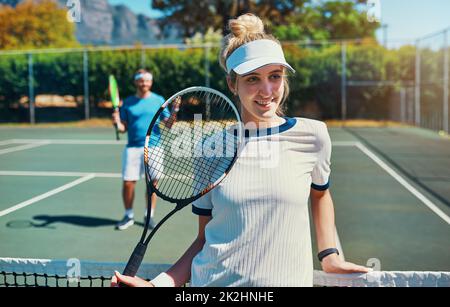 Ich bin immer auf der Siegerseite. Kurze Aufnahme einer attraktiven jungen Tennisspielerin draußen auf dem Platz mit ihrem männlichen Teamkollegen im Hintergrund. Stockfoto