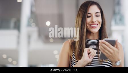 E-Mails unterwegs senden. Ausgeschnittene Aufnahme einer jungen Geschäftsfrau, die ein Smartphone benutzt, während sie durch ein modernes Büro geht. Stockfoto