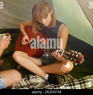 Legen Sie einige Camp Melodien. Aufnahme einer jungen Frau, die in einem Zelt vor ihrem Freund Gitarre spielt. Stockfoto