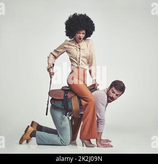 In Bewegung. Eine Studioaufnahme einer attraktiven Frau aus dem Jahr 70s, die einen hübschen Mann mit Sattel auf einer Reitpflanze reitet. Stockfoto