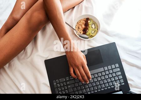 Ich habe diese Frühstücksidee online bekommen. Eine abgeschnittene Aufnahme einer unkenntlichen Frau, die mit ihrem Laptop und einer Haferschale auf ihrem Bett sitzt. Stockfoto