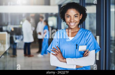 Nichts macht mich glücklicher, als meine Patienten gesund zu sehen. Porträt einer selbstbewussten jungen Ärztin, die im Krankenhaus mit ihren Kollegen im Hintergrund arbeitet. Stockfoto