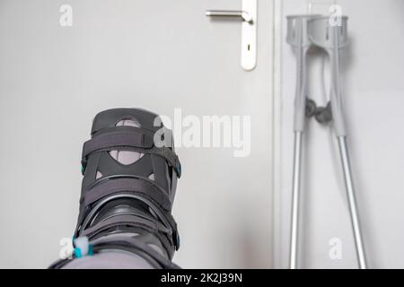 Der europäische Mann nach der Achillessehnenruptur ist wieder zu Hause mit einem speziellen Physiotherapieschuh und Krücken für die Heilung zu Hause mit gesunden Medikamenten Schmerzmittel gegen das schmerzende Bein Stockfoto