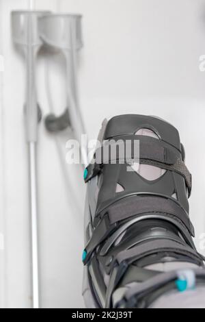 Der europäische Mann nach der Achillessehnenruptur ist wieder zu Hause mit einem speziellen Physiotherapieschuh und Krücken für die Heilung zu Hause mit gesunden Medikamenten Schmerzmittel gegen das schmerzende Bein Stockfoto