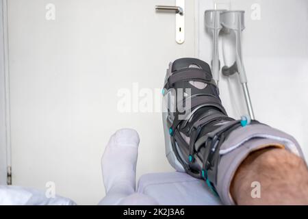 Der europäische Mann nach der Achillessehnenruptur ist wieder zu Hause mit einem speziellen Physiotherapieschuh und Krücken für die Heilung zu Hause mit gesunden Medikamenten Schmerzmittel gegen das schmerzende Bein Stockfoto