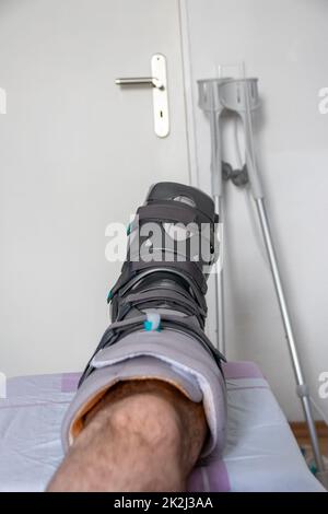 Der europäische Mann nach der Achillessehnenruptur ist wieder zu Hause mit einem speziellen Physiotherapieschuh und Krücken für die Heilung zu Hause mit gesunden Medikamenten Schmerzmittel gegen das schmerzende Bein Stockfoto