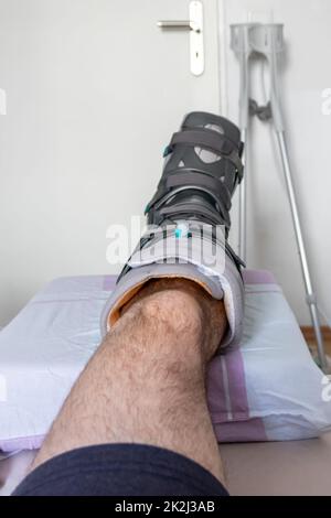 Der europäische Mann nach der Achillessehnenruptur ist wieder zu Hause mit einem speziellen Physiotherapieschuh und Krücken für die Heilung zu Hause mit gesunden Medikamenten Schmerzmittel gegen das schmerzende Bein Stockfoto
