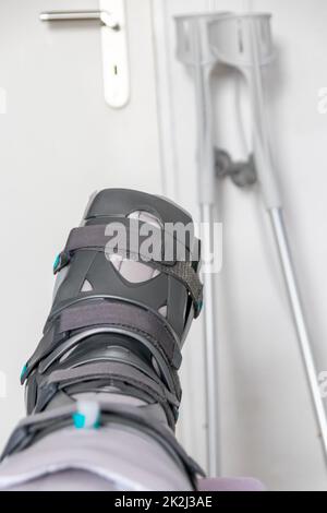 Der europäische Mann nach der Achillessehnenruptur ist wieder zu Hause mit einem speziellen Physiotherapieschuh und Krücken für die Heilung zu Hause mit gesunden Medikamenten Schmerzmittel gegen das schmerzende Bein Stockfoto