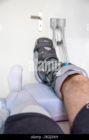 Der europäische Mann nach der Achillessehnenruptur ist wieder zu Hause mit einem speziellen Physiotherapieschuh und Krücken für die Heilung zu Hause mit gesunden Medikamenten Schmerzmittel gegen das schmerzende Bein Stockfoto