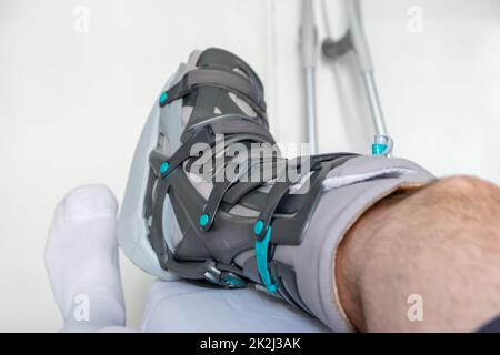 Der europäische Mann nach der Achillessehnenruptur ist wieder zu Hause mit einem speziellen Physiotherapieschuh und Krücken für die Heilung zu Hause mit gesunden Medikamenten Schmerzmittel gegen das schmerzende Bein Stockfoto