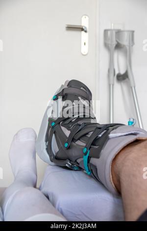 Der europäische Mann nach der Achillessehnenruptur ist wieder zu Hause mit einem speziellen Physiotherapieschuh und Krücken für die Heilung zu Hause mit gesunden Medikamenten Schmerzmittel gegen das schmerzende Bein Stockfoto