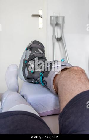 Der europäische Mann nach der Achillessehnenruptur ist wieder zu Hause mit einem speziellen Physiotherapieschuh und Krücken für die Heilung zu Hause mit gesunden Medikamenten Schmerzmittel gegen das schmerzende Bein Stockfoto