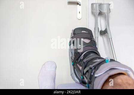 Der europäische Mann nach der Achillessehnenruptur ist wieder zu Hause mit einem speziellen Physiotherapieschuh und Krücken für die Heilung zu Hause mit gesunden Medikamenten Schmerzmittel gegen das schmerzende Bein Stockfoto