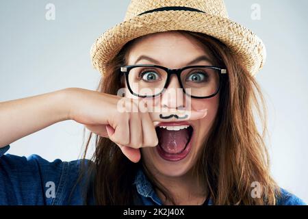 Nehmen Sie das Leben nicht zu ernst. Studioportrait einer jungen Frau, die ihren Finger mit einem Schnurrbart unter die Nase hält. Stockfoto