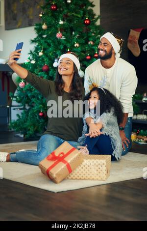 Glückliche Familienerinnerungen wärmen die kalten Dezembertage. Aufnahme einer glücklichen jungen Familie, die Selfies macht, während sie Weihnachtsgeschenke zu Hause eröffnet. Stockfoto