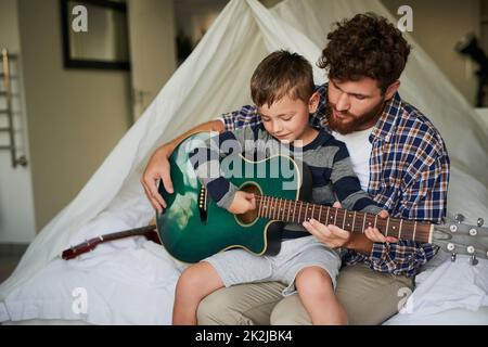 Zeichenfolge für Zeichenfolge. Kurzer Schuss eines entzückenden kleinen Jungen, der auf seinem Schoß seines Vatis sitzt, während er zuhause Gitarre spielen lernt. Stockfoto