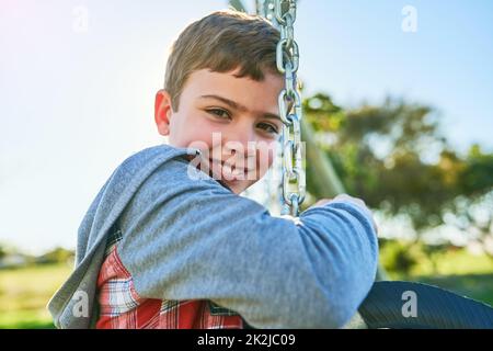 Ich möchte sehen, wie hoch ich schwingen kann. Porträt eines Jungen, der auf einer Schaukel in einem Park sitzt. Stockfoto