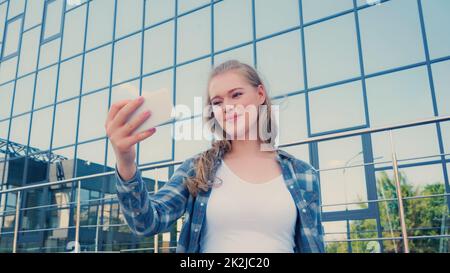 Positive junge Frau im karierten Hemd nimmt Selfie auf dem Smartphone auf der städtischen Straße, Stockbild Stockfoto