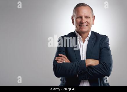 Erfolg war immer das Ziel. Aufnahme eines hübschen, reifen Geschäftsmannes, der mit gefalteten Armen allein vor grauem Hintergrund im Studio steht. Stockfoto