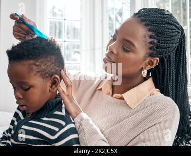 Lass Mama dir helfen. Eine kurze Aufnahme einer attraktiven jungen Frau, die ihrem Sohn die Haare kämmt, während sie zu Hause auf einem Bett sitzt. Stockfoto