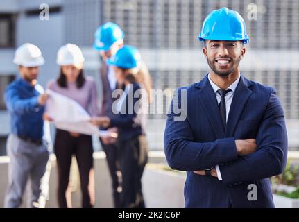 Ich habe das Team, das es möglich machen kann. Foto eines jungen Geschäftsmannes, der mit zusammengefalteten Armen steht und einen Schutzhelm trägt, während seine Kollegen hinter ihm stehen. Stockfoto