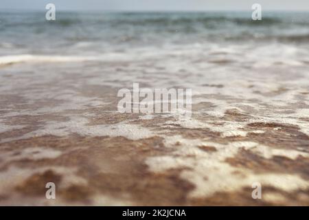 Flachwinkelfoto vom Erdgeschoss - Strand mit flachem Wasser und weißem Schaum über Sand, nasse Lufttropfen, verschwommenes Meer in der Ferne. Abstrakter Urlaubshintergrund. Stockfoto