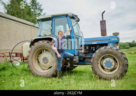 Kommen Sie und schauen Sie sich in mein Büro. Porträt eines fröhlichen jungen Bauern, der neben seinem großen Traktor auf einem Feld posiert. Stockfoto