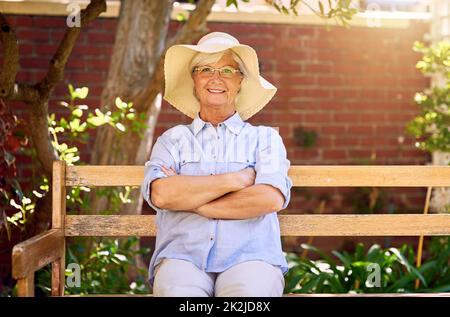 Im Garten zu sein bringt mir ein Lächeln ins Gesicht. Porträt einer glücklichen älteren Frau, die sich auf einer Bank im Garten entspannt. Stockfoto