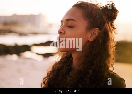 Das Universum spricht, wenn man anhält und zuhört. Aufnahme einer attraktiven jungen Frau, die allein auf einer Matte sitzt und bei Sonnenuntergang am Strand meditiert. Stockfoto