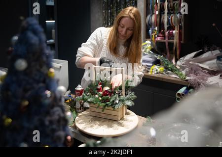 Minsk, Weißrussland - 20. Dez 2021: Ein Mitarbeiter eines Blumenbodens kreiert ein Blumenarrangement in einer weihnachtlichen Holzkiste Stockfoto