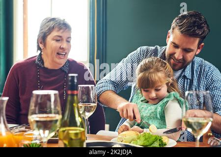 Lasst mich euch dort helfen, liebe. Aufnahme eines fröhlichen jungen Mannes, der seinen Töchtern beim Essen half, während er zu Hause an einem Esstisch saß. Stockfoto