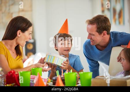 Ich glaube, ich weiß, was das ist. Aufnahme eines kleinen Jungen, der seine Geburtstagsgeschenke inmitten seiner Familie öffnete. Stockfoto