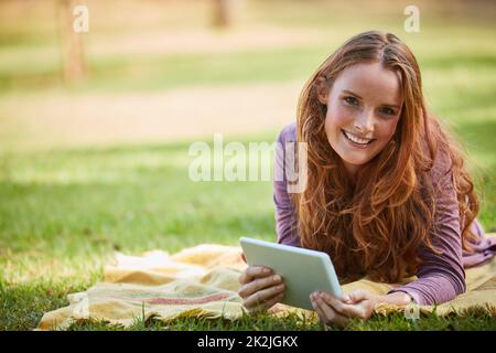 Ich kann mir keinen besseren Weg vorstellen, den Tag zu verbringen. Aufnahme einer jungen Frau, die ihr Tablet benutzt, während sie sich im Park entspannt. Stockfoto