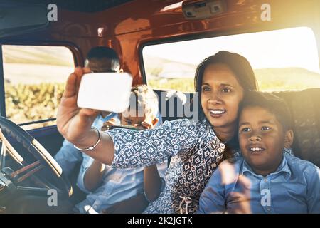 Zeit für ein paar Fotos für die Erinnerungen. Aufnahme einer fröhlichen jungen Familie, die in einem roten Pick-up auf einer Landstraße fährt und ein Selbstporträt auf einem Handy zusammen macht. Stockfoto