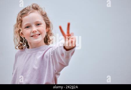 Ein selbstbewusstes Kind ist ein glückliches Kind. Studioaufnahme eines kleinen Mädchens, das vor grauem Hintergrund ein Friedenszeichen macht. Stockfoto