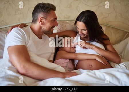 Und bald werden es vier sein. Aufnahme eines glücklichen kleinen Mädchens, das mit ihrem Vater und ihrer Schwangeren im Bett liegt. Stockfoto